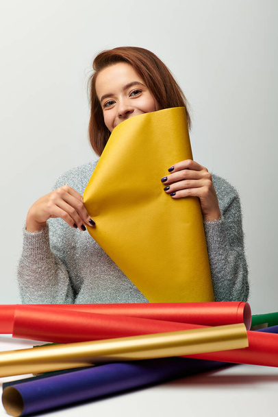 season of joy, young woman in sweater holding yellow Christmas gift paper on grey backdrop - Photo, Image