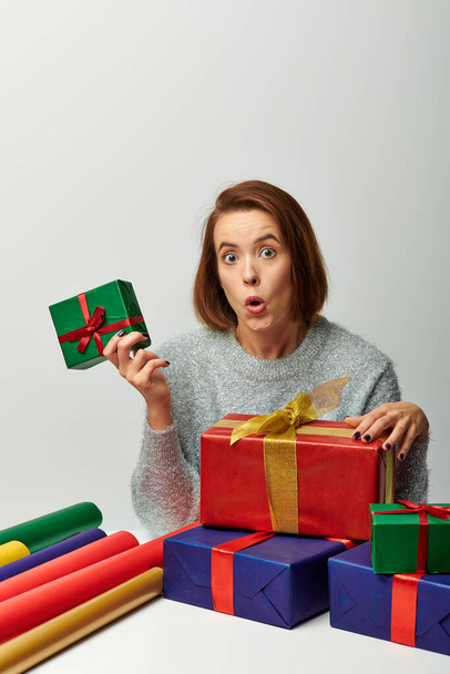 Wow-Gefühl, Frau im Winterpullover hält Weihnachtsgeschenk in der Nähe von buntem Geschenkpapier auf grau - Foto, Bild