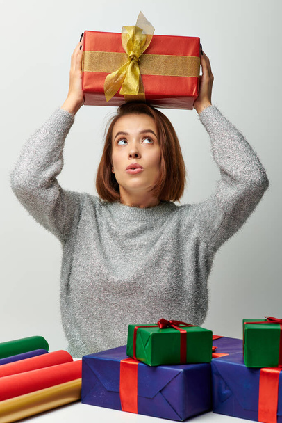 thrilled woman in winter sweater holding Christmas present above head near gift paper on grey - Photo, Image