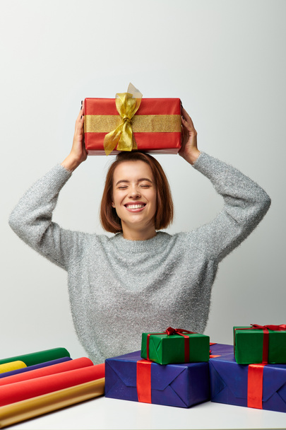 Frau im Winterpullover hält Weihnachtsgeschenk über Kopf in der Nähe von Geschenkpapier auf grau - Foto, Bild