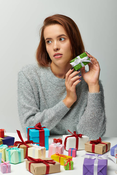 schöne Frau in kuscheligem Pullover mit winzigem Weihnachtsgeschenk in der Nähe bunt verpackter Geschenke auf grau - Foto, Bild