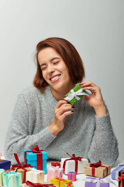 wesoła kobieta w przytulnym swetrze trzymając malutki prezent świąteczny w pobliżu kolorowych zapakowanych prezentów na szaro - Zdjęcie, obraz