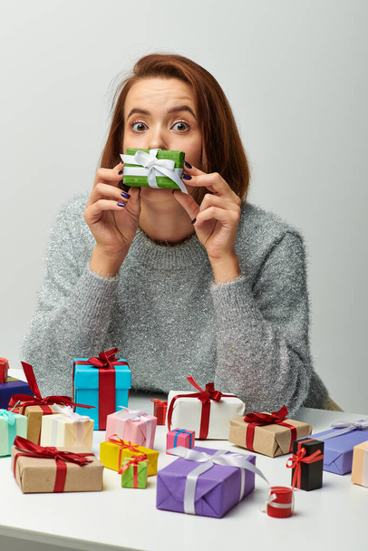 смішна жінка в светрі тримає крихітний різдвяний подарунок біля обличчя і барвисті загорнуті подарунки на сірому - Фото, зображення