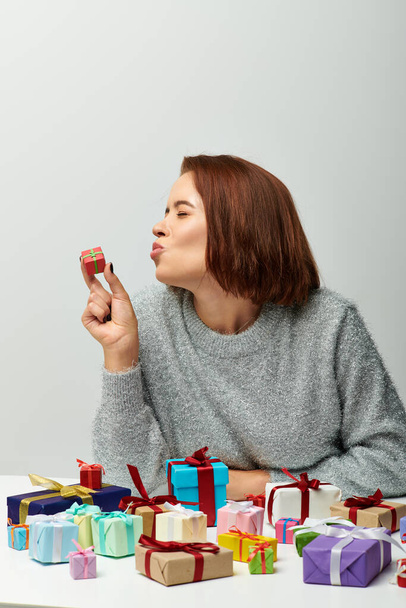 young woman in cozy sweater kissing tiny Christmas present near colorful wrapped gifts on grey - Photo, Image