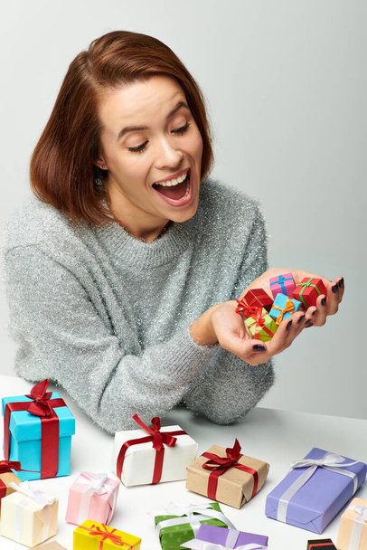 カラフルな包まれた贈り物の近くに小さなクリスマスプレゼントの束を見ている居心地の良いセーターに興奮した女性 - 写真・画像