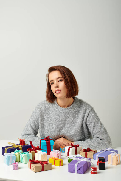 attractive woman in sweater sitting among bunch of Christmas presents on table, grey backdrop - Photo, Image