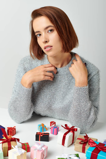 beautiful woman in winter sweater sitting among bunch of Christmas presents on table, grey backdrop - Photo, Image