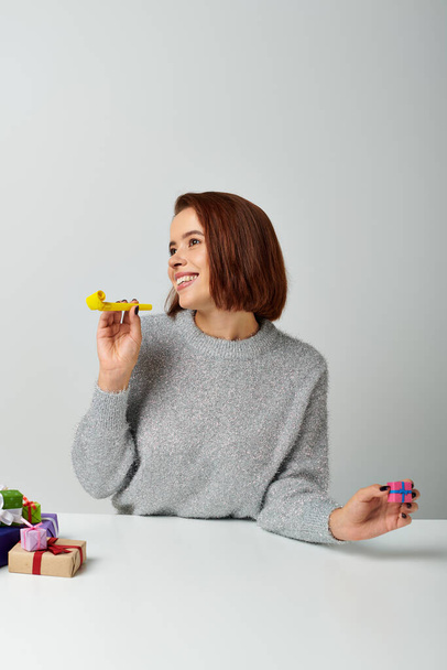 cheerful woman in sweater fantasizing and holding tiny Christmas gift and yellow party horn on grey - Photo, Image