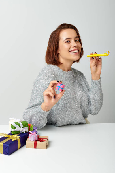 happy woman in sweater smiling and holding tiny Christmas gift and yellow party horn on grey - Photo, Image