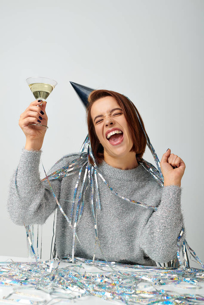 excited woman in party cap with tinsel on head holding glass of champagne on grey, Happy New Year - Photo, Image