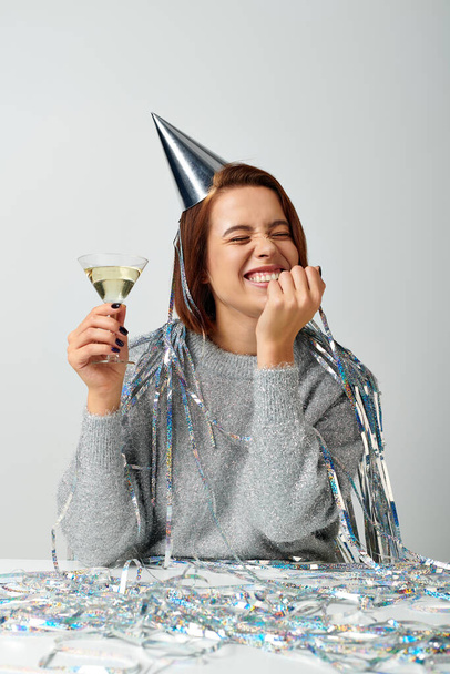 thrilled woman in party cap with tinsel on head holding glass of champagne on grey, Happy New Year - Photo, Image