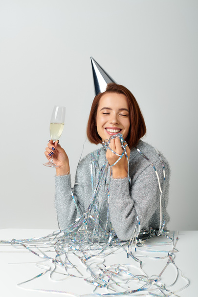 glückliche Frau mit Partymütze und Lametta auf dem Kopf, die ein Champagnerglas hält, während sie einen Neujahrswunsch äußert - Foto, Bild