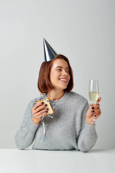 szczęśliwa kobieta w party cap trzyma kanapkę z świecidełkiem i kieliszkiem szampana, świętując Nowy Rok - Zdjęcie, obraz