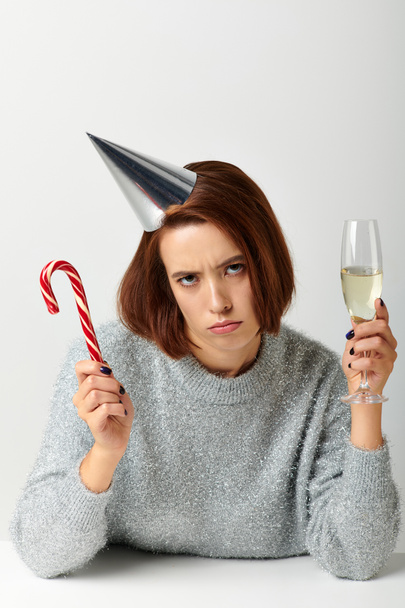 mujer disgustada en la tapa del partido sosteniendo copa de champán y bastón de caramelo en gris, Feliz Navidad - Foto, imagen