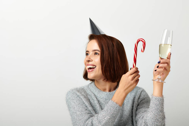mujer alegre en la tapa del partido sosteniendo copa de champán y bastón de caramelo en gris, concepto de Feliz Navidad - Foto, Imagen