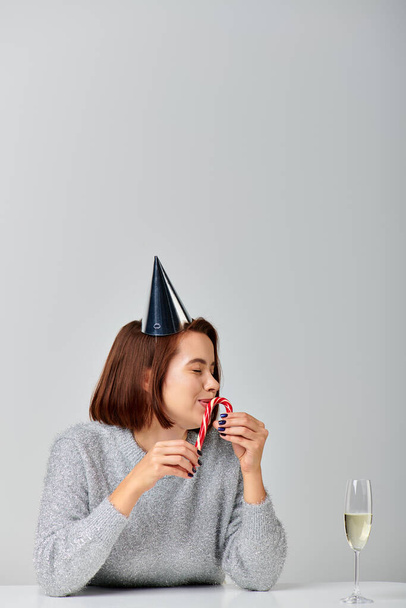mulher feliz no tampão do partido beijando cana-de-açúcar perto de copo de champanhe no fundo cinza, Feliz Natal - Foto, Imagem