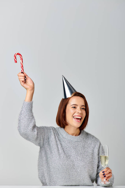 femme excitée dans le chapeau de fête tenant canne à bonbons et verre de champagne sur fond gris, Joyeux Noël - Photo, image