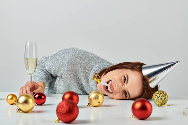 Женщина в кепке для вечеринок лежит на столе возле бокала шампанского и рождественских безделушек, трубит в рог вечеринки - Фото, изображение