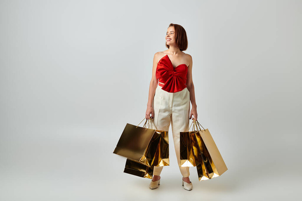 διακοπές ψώνια, ευτυχισμένη γυναίκα σε μοντέρνα ενδυμασία κρατώντας τσάντες ψώνια σε γκρι φόντο, Χριστούγεννα - Φωτογραφία, εικόνα