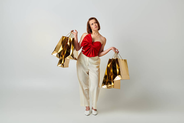 holiday shopping, confused woman in stylish attire holding shopping bags on grey background - Photo, Image