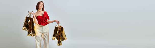 banner de compras navideñas, mujer pensativa con un atuendo elegante sosteniendo bolsas de compras sobre fondo gris - Foto, Imagen