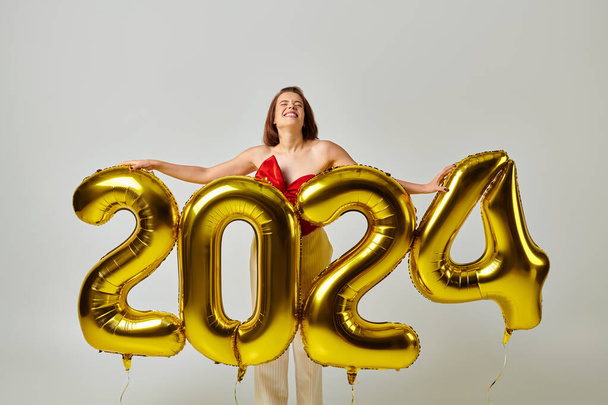 Ευτυχισμένο το νέο έτος, χαρούμενη νεαρή γυναίκα σε μοντέρνα ενδυμασία κρατώντας μπαλόνια με 2024 αριθμούς σε γκρι - Φωτογραφία, εικόνα