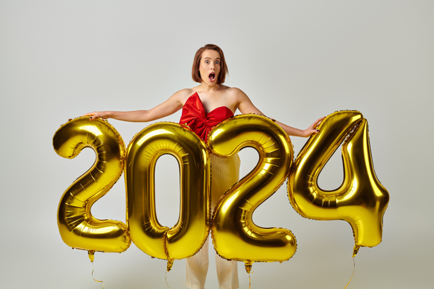 Bonne année, choquée jeune femme en tenue tendance tenant des ballons avec des numéros 2024 sur gris - Photo, image