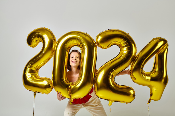 Bonne année, jeune femme heureuse en tenue tendance tenant des ballons avec des numéros 2024 sur gris - Photo, image