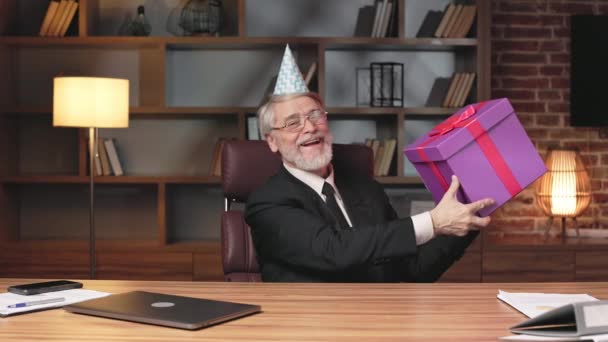 Well-dressed aged gentleman in party hat demonstrating present while sitting in executive chair of modern office. Happy business person rejoicing anniversary of company success in cozy workplace. - Footage, Video