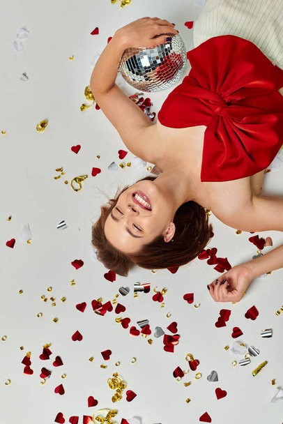 Новый год, возбужденная женщина с дискотечным мячом, лежащая на полу рядом с конфетти на сером заднем плане, вид сверху - Фото, изображение