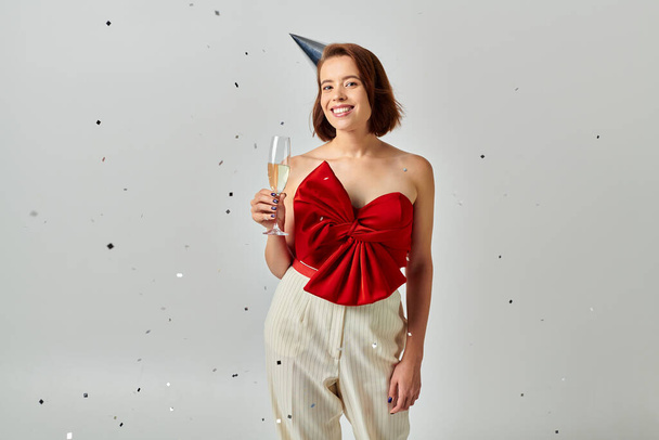Feliz Año Nuevo, mujer alegre con gorra de fiesta sosteniendo copa de champán cerca de confeti en gris - Foto, Imagen