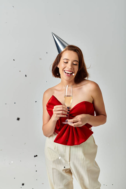 Feliz Año Nuevo, mujer joven positiva en la tapa del partido sosteniendo la copa de champán cerca de confeti en gris - Foto, Imagen