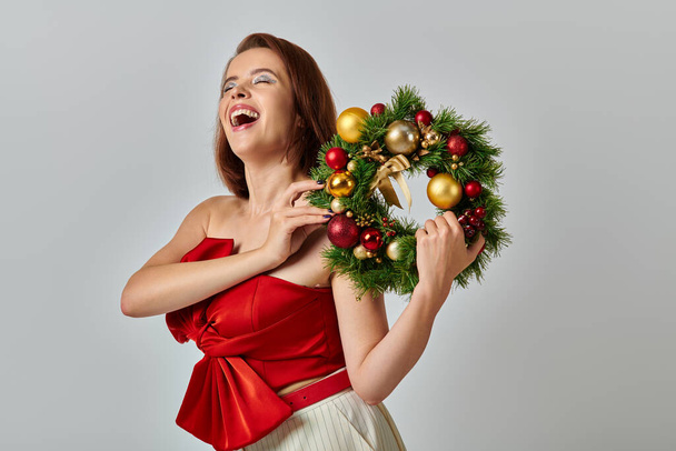 świąteczny sezon, inspirowana kobieta z błyszczącym makijażem trzymając świąteczny wieniec na szarym tle - Zdjęcie, obraz
