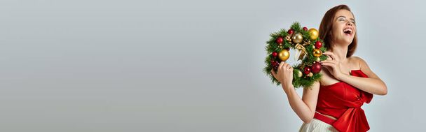 świąteczny sztandar sezonu, podekscytowana kobieta z błyszczącym makijażem trzymając świąteczny wieniec na szarym tle - Zdjęcie, obraz