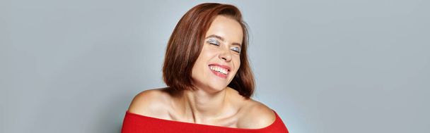 zadowolona młoda kobieta z brunetką krótkie włosy pozowanie w czerwonym stroju na szarym tle, baner - Zdjęcie, obraz