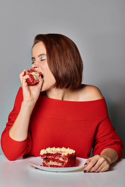młoda kobieta w czerwonym stroju gryzie pyszny kawałek ciasta bento na szarym tle, wszystkiego najlepszego z okazji urodzin - Zdjęcie, obraz