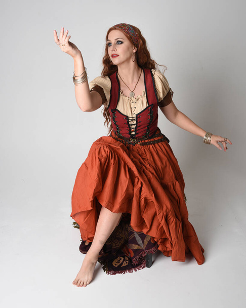 Retrato de cuerpo entero de una hermosa mujer pelirroja vestida con una doncella medieval, disfraz de adivina. Posar sentado, con las manos gestuales extendidas. aislado en estudio  - Foto, imagen
