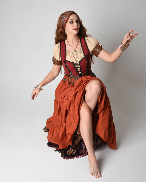 Volledig portret van een prachtige roodharige vrouw in een middeleeuws meisjeskostuum. Zittende houding, met gebarentanden die reiken. geïsoleerd op studio  - Foto, afbeelding