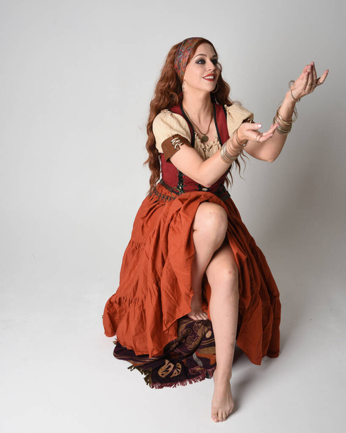 Ritratto a figura intera di bella donna dai capelli rossi con indosso una fanciulla medievale, costume da indovino. Posizione seduta, con le mani gestuali tese verso l'esterno. isolato in studio  - Foto, immagini