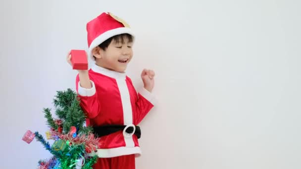 Ázsiai fiú télapó jelmezt visel Állva és játszik közel egy mókás karácsonyfa. 4K felbontás. - Felvétel, videó