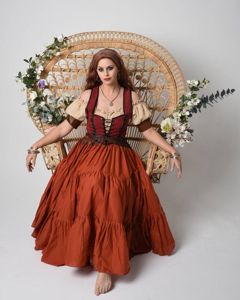 Volledig portret van een prachtige roodharige vrouw in een middeleeuws meisjeskostuum. Zittende houding, met gebarentanden die reiken. geïsoleerd op studio achtergrond. - Foto, afbeelding