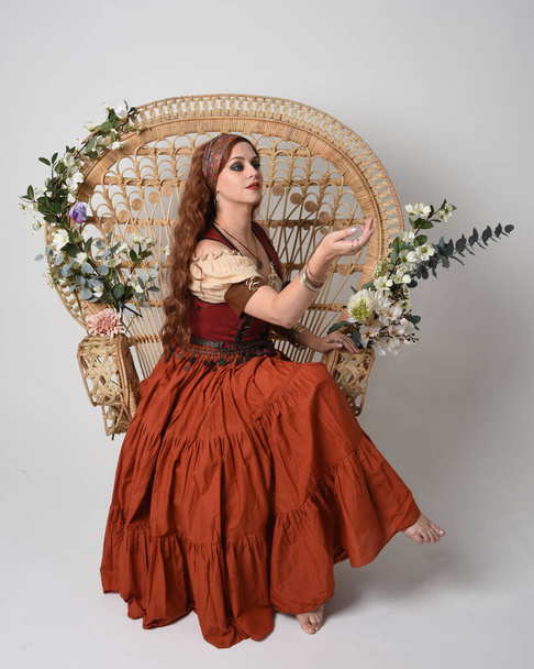 Full length portrait of beautiful red haired woman wearing a medieval maiden, fortune teller costume.  Sitting pose, with gestural hands reaching out. isolated on studio background. - Photo, Image