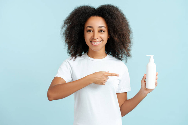 Portré mosolygós afro-amerikai nő göndör haj fehér pólóban kezében palack mockup ujjal mutogatott kamera elszigetelt kék háttér mockup. Fogalom a reklám, hajápolás - Fotó, kép