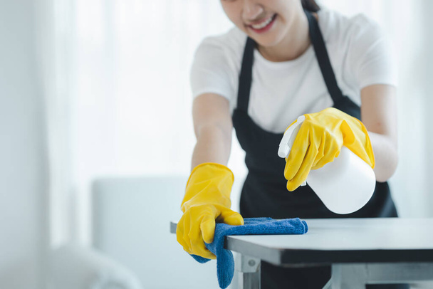 Asian female cleaner wiping down tables with cleaning spray, Wear rubber gloves and an apron and work with a happy smile, Use a towel to wipe the table, cleaning idea. - Photo, Image