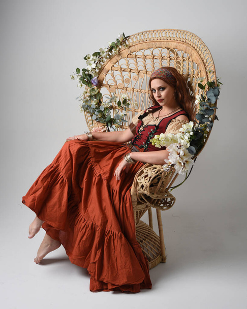 Portret pięknej rudej kobiety w średniowiecznym kostiumie wróżki. Siedząca poza, z gestywnymi dłońmi wyciągniętymi do góry. izolowany na tle studia. - Zdjęcie, obraz