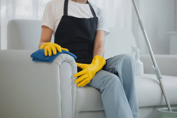Personel sprzątający sprząta meble wewnątrz domu, nosić gumowe rękawiczki i fartuch i pracować z uśmiechem szczęśliwy, Użyj ręcznika, aby wytrzeć kanapę, pomysł czyszczenia. - Zdjęcie, obraz