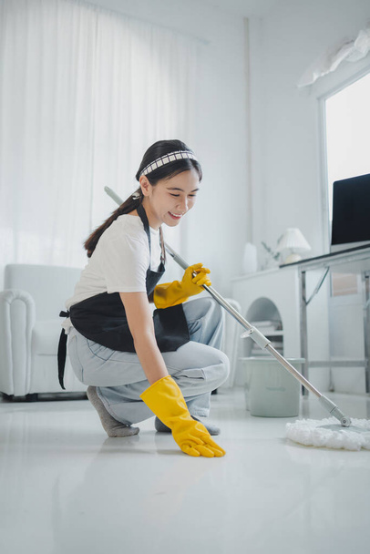 Het schoonmaakpersoneel controleert de vloer om te zien of het schoon is of niet, draagt rubberen handschoenen en een schort en werkt met een vrolijke glimlach, werkt met schoonmaakmiddelen, schoonmaakidee. - Foto, afbeelding