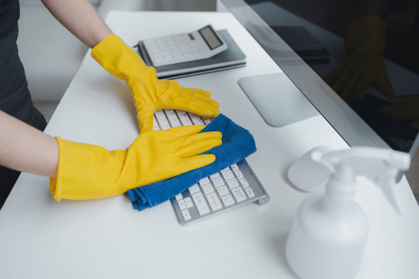 Reinigungspersonal wischt Bürogeräte ab, wischt die Tastatur mit einem Handtuch und Desinfektionsmittel sauber. Tragen Sie Gummihandschuhe bei der Arbeit mit Reinigungschemikalien, Reinigungsidee. - Foto, Bild
