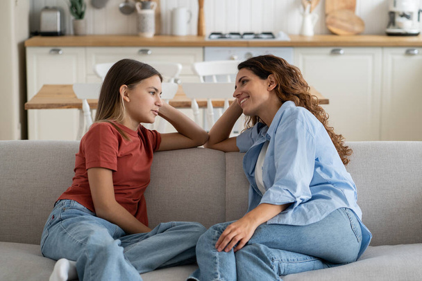 Happy charismatic Italian woman talking with daughter about school friends and summer vacation plans sits on sofa in home interior. Shy casual teen girl shares secrets with mom thanks to family trust  - Photo, Image