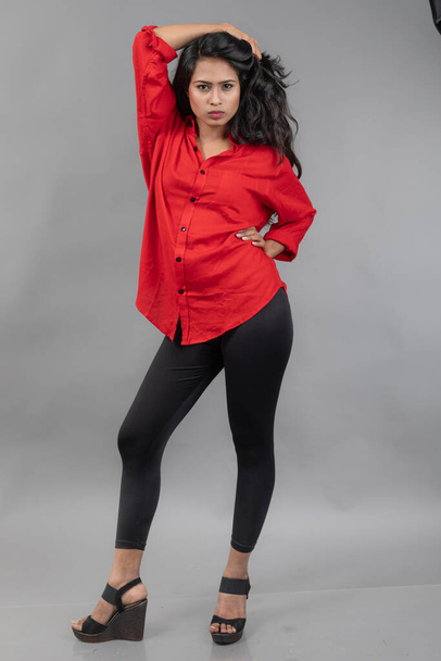 Индийская молодая модель в повседневной одежде на сером фоне - фото на складе. Длинные черные волосы модели в красной рубашке и черных джинсах. - Фото, изображение